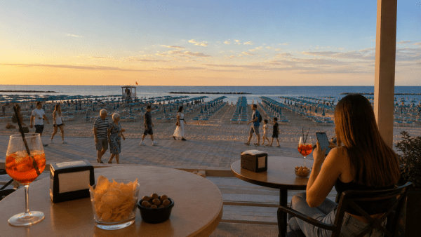 Un tavolino di un bar sul lungomare romagnolo al tramonto. In primo piano uno Spritz, sul tavolo accanto una ragazza osservata di spalle mentre scorre lo smartphone. Sullo sfondo la passeggiata, la spiaggia e una coppia di anziani che cammina lentamente.