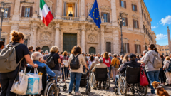 Manifestazione di caregiver e persone con disabilità davanti a Montecitorio, a Roma. Una folla con carrozzine, deambulatori, medicinali e borse per l'assistenza quotidiana si raccoglie davanti al Parlamento, mentre una figura anonima osserva dall'alto.
